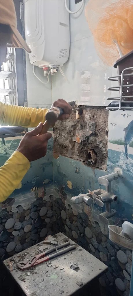 Worker chiselling a bathroom wall open around a geyser inlet to reach the leaking pipe